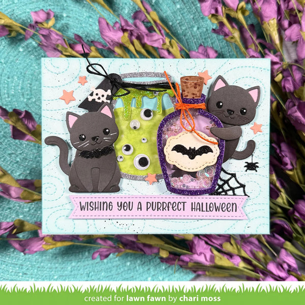 A handmade Halloween greeting card featuring two cartoon gray cats, a green slime jar, a purple potion bottle, and a pink banner reading 'Wishing you a Purrfect Halloween.' The card is displayed on a turquoise surface surrounded by purple flowers.