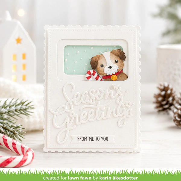 A handmade greeting card with a white embossed border and the words 'Seasons Greetings' in cursive, featuring a small illustrated dog with a red-and-white striped scarf and the text 'FROM ME TO YOU' at the bottom. The card is styled on a light surface with holiday props including candy canes, pine branches, a house ornament, and pinecones.