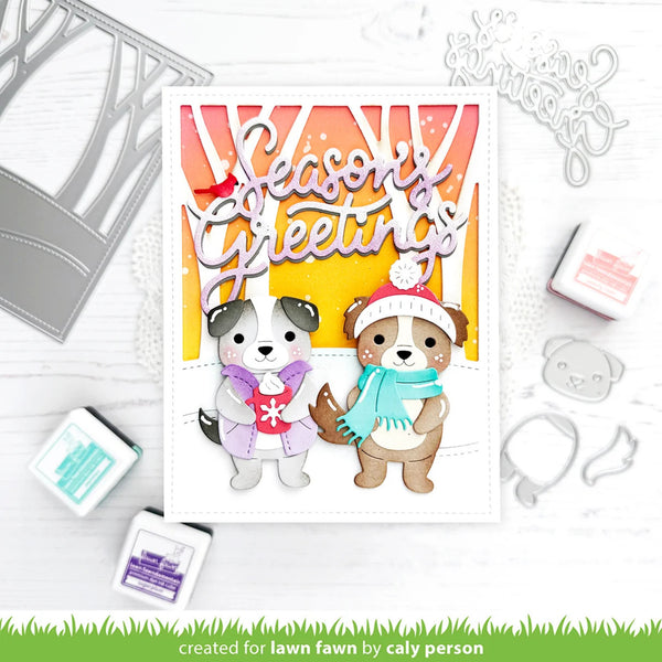 A handmade holiday greeting card featuring two cartoon dogs in winter clothing under a colorful striped arch with the words 'Season's Greetings.' The card is styled on a white surface with die-cut embellishments, stamps, and ink pads around it.