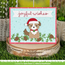 A handmade Christmas greeting card featuring a cute brown dog wearing a red Santa hat, set against a light blue background with pine branches and the words 'joyful wishes' in red script. The card is displayed on a rustic birch log slice with artificial grass and cacti in the background.
