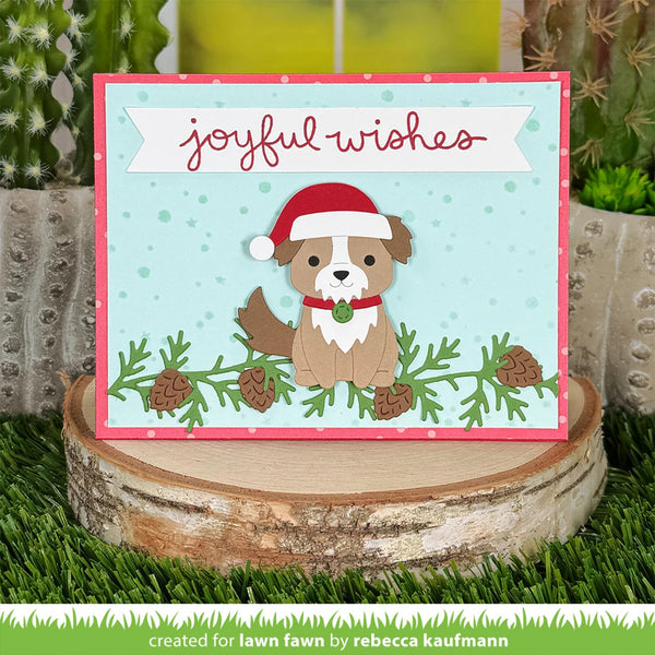 A handmade Christmas greeting card featuring a cute brown dog wearing a red Santa hat, set against a light blue background with pine branches and the words 'joyful wishes' in red script. The card is displayed on a rustic birch log slice with artificial grass and cacti in the background.