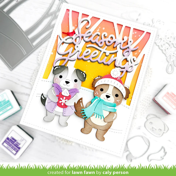 A handmade layered card featuring two cute cartoon dogs wearing winter hats and scarves under a 'Season's Greetings' banner. The card is styled on a white surface with crafting tools, stamps, and ink pads around it.