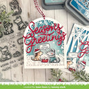 A handmade Christmas card featuring layered die-cut paper with a winter dog illustration, red cursive 'Seasons Greetings' text, and a blue snowflake-patterned background. The card is styled on a whitewashed wood surface with clear stamped designs, ink pads, and additional card elements.