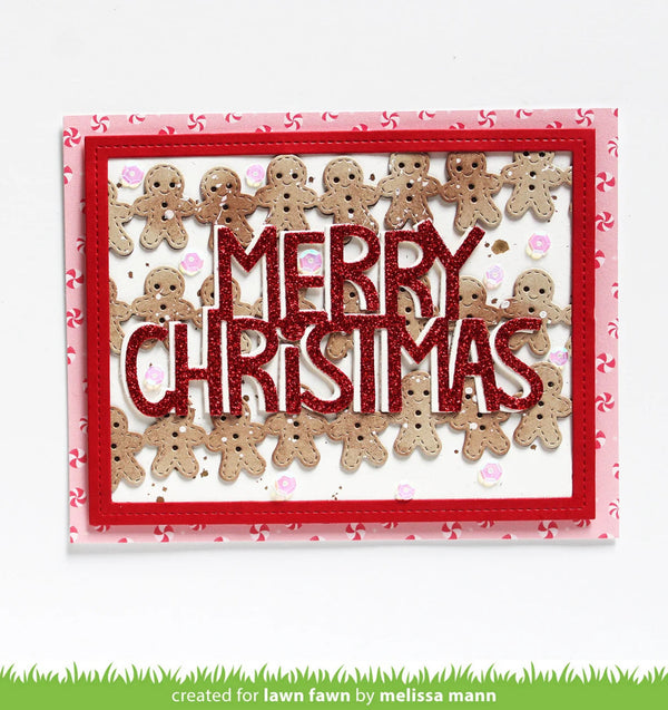 A handmade Christmas card featuring the words 'MERRY CHRISTMAS' in glittery red letters, surrounded by small gingerbread men figures. The card has a red border and a pink patterned backing, shown against a white background with a green grass graphic at the bottom.