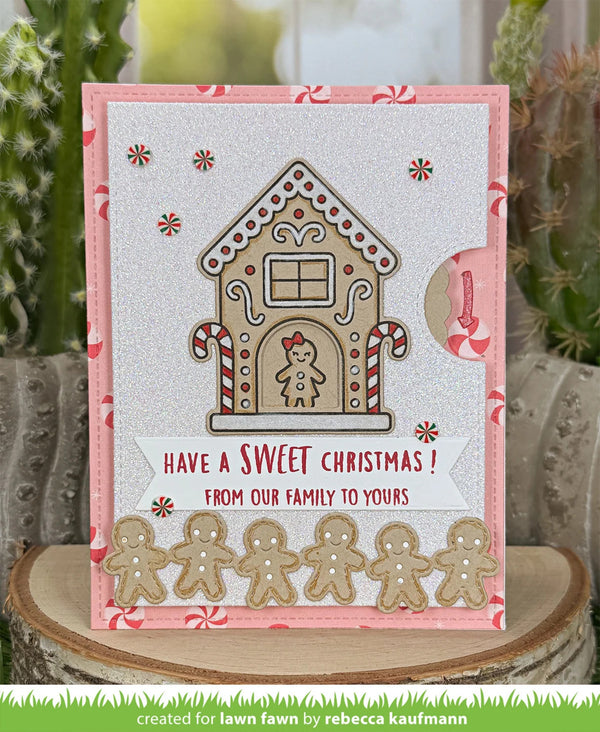 A handmade Christmas greeting card featuring a gingerbread house design, candy canes, and gingerbread men cutouts, displayed on a wooden slice with cacti in the background.