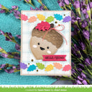 A handmade greeting card featuring a cute acorn face with a smiling expression, decorated with colorful autumn leaves and the text 'HELLO, FRIEND'. The card is displayed on a textured turquoise surface surrounded by purple flowers.