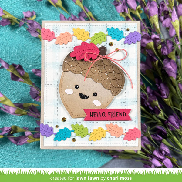 A handmade greeting card featuring a cute acorn face with a smiling expression, decorated with colorful autumn leaves and the text 'HELLO, FRIEND'. The card is displayed on a textured turquoise surface surrounded by purple flowers.