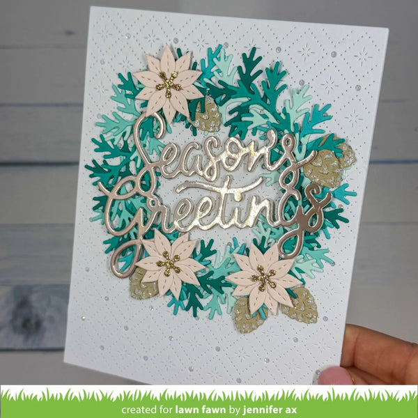 A handmade holiday greeting card held by a hand, featuring layered die-cut paper snowflakes and poinsettias in teal, gold, and cream, with a central metallic script reading 'Season's Greetings'.