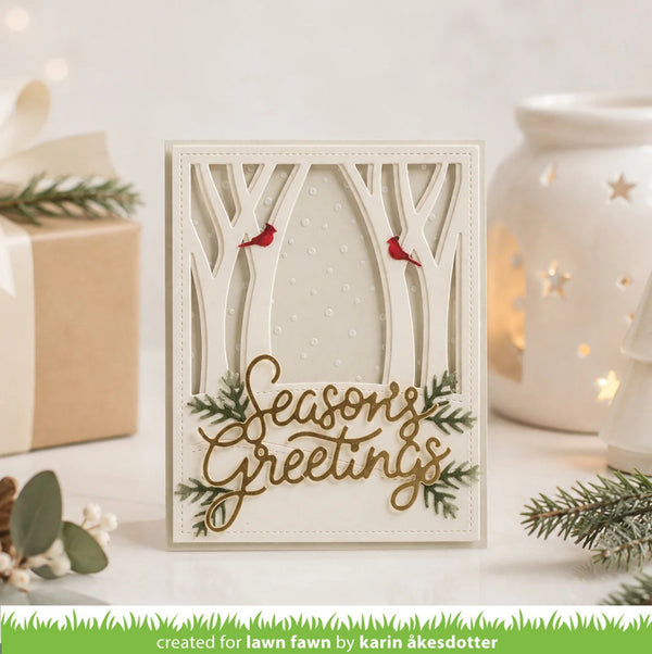 A handmade layered card with a forest theme, featuring laser-cut tree branches, two small red birds, green pine branches, and the words 'Seasons Greetings' in gold. The card is displayed on a white surface with a warm white ceramic candle holder and a kraft gift box in the background.