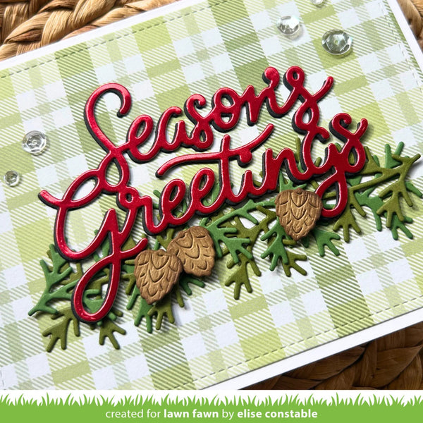 A close-up of a handmade holiday greeting card featuring the words 'Seasons Greetings' in red script over a green and white gingham background, with layered green pine branches and two small pinecones. A green banner at the bottom reads 'created for lawn fawn by elise constable'.