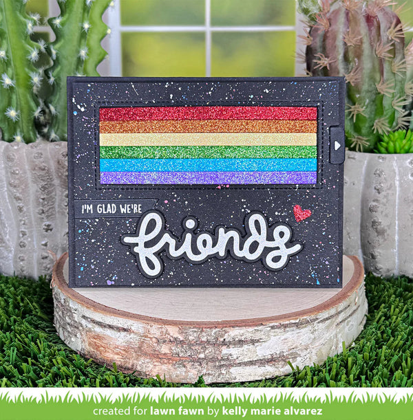 Card with rainbow design and 'I'm glad we're friends' text, placed on a wooden stump with cacti in the background.