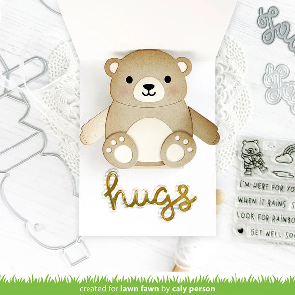 A handmade greeting card featuring a cute layered teddy bear with a gold foil 'hugs' script on a white background, surrounded by crafting tools and a clear stamp sheet.