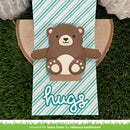 A handmade paper craft card featuring a cute brown bear with a cream snout and paws, set against a mint green and white striped background with the word 'hugs' in white script. The card is displayed on a green artificial grass background with decorative lace flowers.