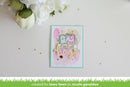 A handmade greeting card featuring two cute cartoon characters with a pink and gold 'Sending Love' banner, styled on a white surface with white flowers in the corner. A green banner at the bottom reads 'created for lawn fawn by nicole gavaldon'.