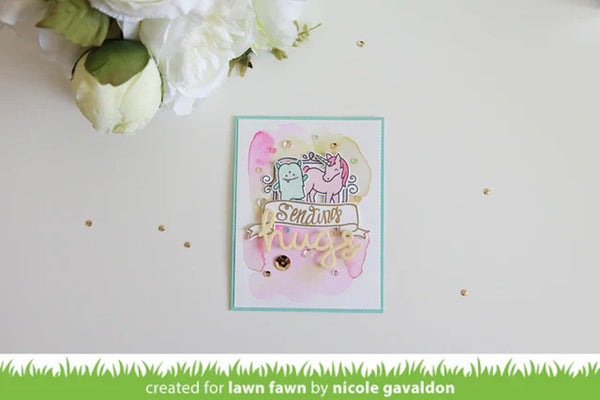A handmade greeting card featuring two cute cartoon characters with a pink and gold 'Sending Love' banner, styled on a white surface with white flowers in the corner. A green banner at the bottom reads 'created for lawn fawn by nicole gavaldon'.