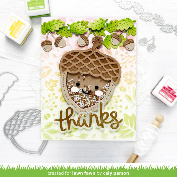 A handmade autumn-themed card featuring a large layered acorn with a smiling face and the word 'thanks' in metallic gold script. The card is styled on a white surface with craft tools, stamps, and ink pads around it.