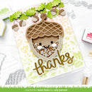 A handmade autumn-themed card featuring a cute acorn face with a smiling face peeking out from inside, decorated with green leaves and acorns along the top. The card has a gold 'thanks' script at the bottom and is displayed on a white background with crafting tools.