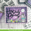 A Halloween-themed handmade card featuring the words 'Hocus Pocus' in large black script over a purple starry background, with illustrated cats in witch hats, potion bottles, a broom, books, and a speech bubble reading 'You're Spell Call to Me.' The card is photographed on a white wood surface with crafting supplies around it, including clear stamps, purple thread, and ink pads.