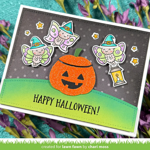 A handmade Halloween greeting card with a glittery orange jack-o'-lantern, three cute fairy cutouts, and the words “HAPPY HALLOWEEN!” on a dark gray and green background, styled with purple flowers.