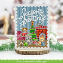 A handmade holiday greeting card featuring cartoon dogs around a Christmas tree and fireplace, displayed on a wood slice stand with blurred festive greenery in the background.