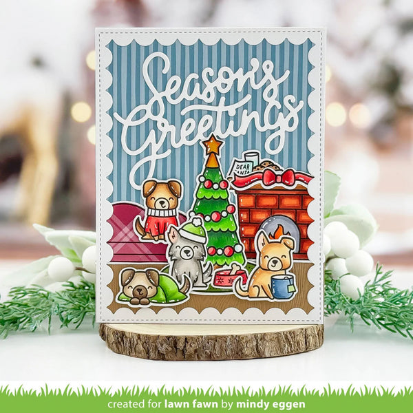 A handmade holiday greeting card featuring cartoon dogs around a Christmas tree and fireplace, displayed on a wood slice stand with blurred festive greenery in the background.