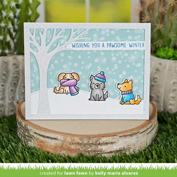 A handmade greeting card with a winter scene featuring three cute dogs, displayed on a rustic wood slice with artificial grass and plants in the background. The card reads 'WISHING YOU A PAWSOME WINTER' and is created for lawn fawn by Kelly Marie Alvarez.
