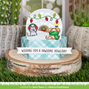 A handmade greeting card or gift box set displayed on a wood slice with artificial grass and potted cacti. The central item is a round white card featuring two dogs in winter clothing under holiday lights, with a blue-and-white checkered base and a white banner reading 'WISHING YOU A PAWSOME HOWIDAY.