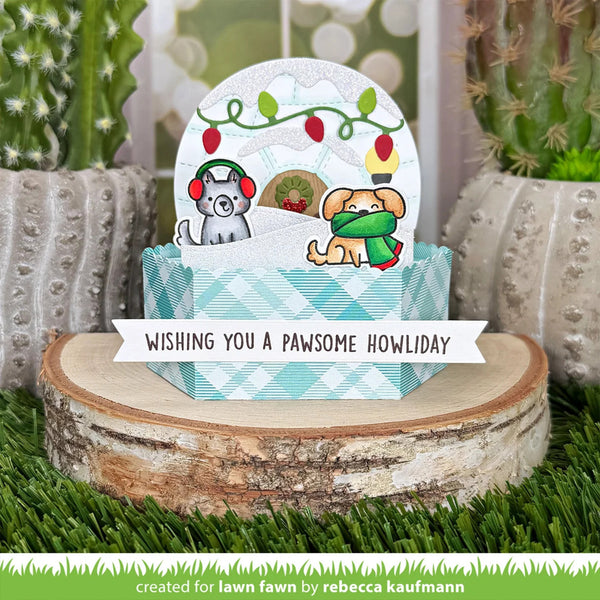 A handmade greeting card or gift box set displayed on a wood slice with artificial grass and potted cacti. The central item is a round white card featuring two dogs in winter clothing under holiday lights, with a blue-and-white checkered base and a white banner reading 'WISHING YOU A PAWSOME HOWIDAY.