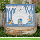 A handmade winter-themed greeting card displayed on a rustic wood slice stand. The card features a blue textured border, a snowy igloo scene with bare tree silhouettes, three cartoon dogs wearing blue hats and sweaters, and the text 'WISHING YOU A COZY WINTER' and 'HAPPY HOLIDAYS'.