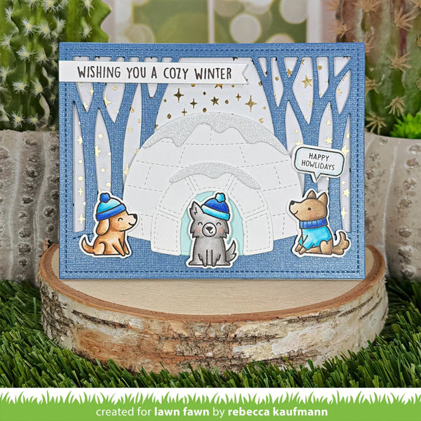 A handmade winter-themed greeting card displayed on a rustic wood slice stand. The card features a blue textured border, a snowy igloo scene with bare tree silhouettes, three cartoon dogs wearing blue hats and sweaters, and the text 'WISHING YOU A COZY WINTER' and 'HAPPY HOLIDAYS'.