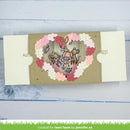 A handmade layered paper craft card featuring a heart-shaped window with a squirrel and two monkeys inside, surrounded by a floral leaf border in pink, red, and cream. The card is displayed on a light blue wooden surface.