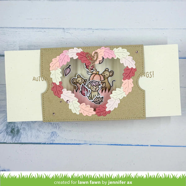 A handmade layered paper craft card featuring a heart-shaped window with a squirrel and two monkeys inside, surrounded by a floral leaf border in pink, red, and cream. The card is displayed on a light blue wooden surface.