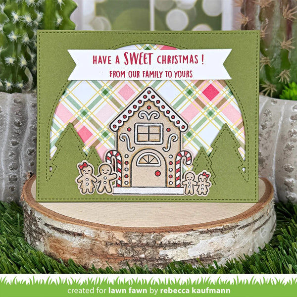 A handmade Christmas greeting card displayed on a rustic wood slice with greenery in the background. The card features an olive green base, a gingerbread house design, holiday plaid accents, and a white banner reading 'HAVE A SWEET CHRISTMAS! FROM OUR FAMILY TO YOURS'.