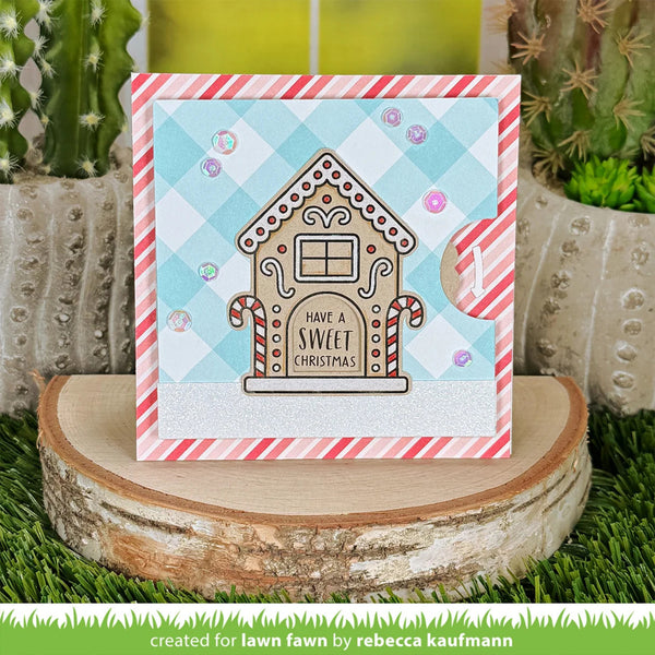A handmade Christmas greeting card featuring a gingerbread house with the text 'HAVE A SWEET CHRISTMAS' on a light blue and white gingham background, styled on a wood slice with cacti in the background.
