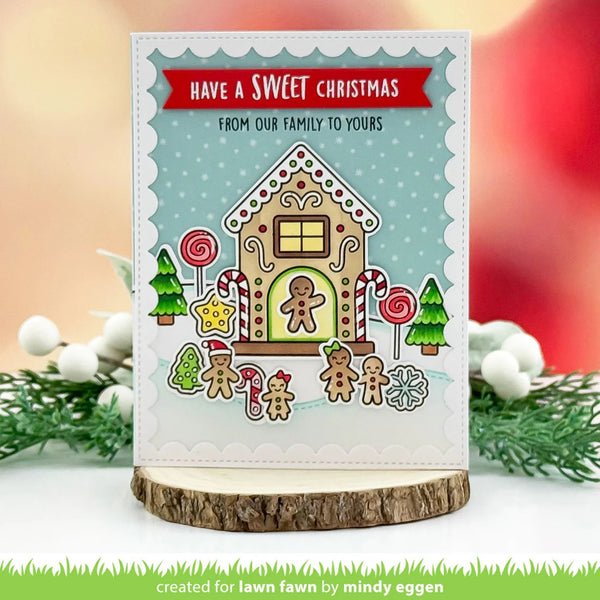 A handmade Christmas greeting card featuring a gingerbread house illustration with the text 'HAVE A SWEET CHRISTMAS FROM OUR FAMILY TO YOURS' on a scalloped white border. The card is displayed on a wood slice stand with blurred holiday greenery and red background bokeh.