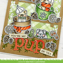 A close-up of a handmade holiday-themed card featuring two cute illustrated dogs in teacups, pinecones, snowflakes, and the words 'YOU'RE MY pup OF TEA'. The card is photographed on a white wood surface, with a green grass-like banner at the bottom reading 'created for lawn fawn by june helsen'.