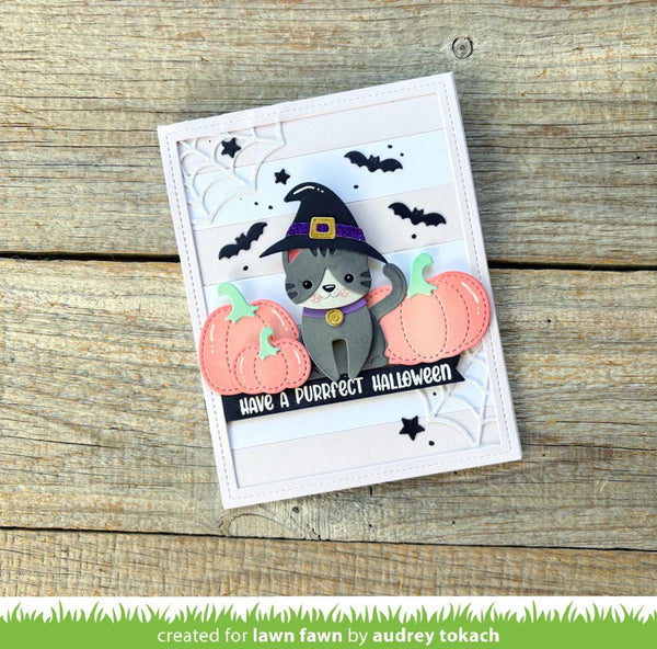 A handmade Halloween greeting card featuring a gray cat in a witch hat with two pink pumpkins and black bats, placed on a rustic wooden surface.