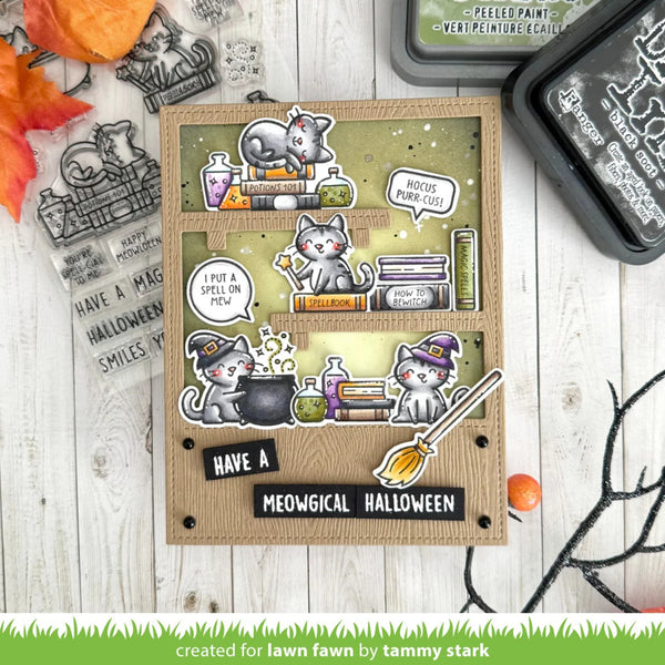 A handmade scrapbook card featuring three illustrated cats in witch hats around cauldrons and books, styled on a white wood background with Halloween-themed props and stamp blocks.
