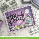 A handmade Halloween-themed card with a purple and lavender background, featuring the words 'Hocus Pocus' in large black script, a white cat dressed as a witch sitting on books, potion bottles, and a speech bubble that says 'YOU'RE SPELLICAL TO ME.' The card is styled on a light wood surface with clear stamps, ink pads, and craft supplies around it.