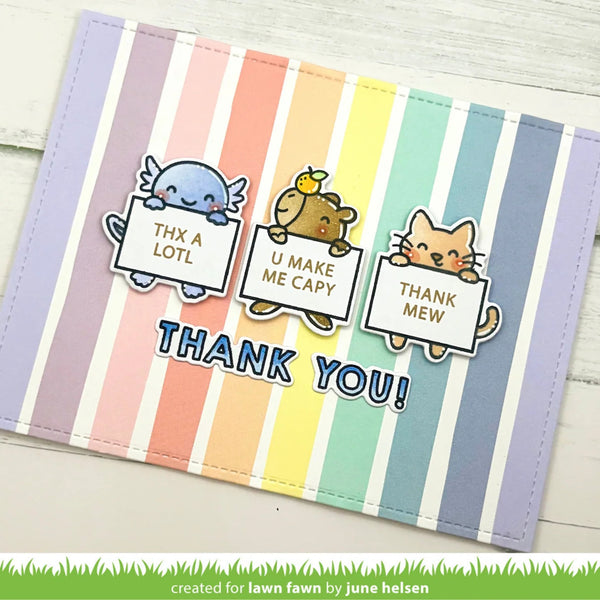 A handmade greeting card with pastel rainbow vertical stripes and cute cartoon animals holding signs that read 'THX A LOTL', 'U MAKE ME CAPY', and 'THANK MEW', with 'THANK YOU!' in blue letters across the bottom. The card is photographed on a light wood surface.