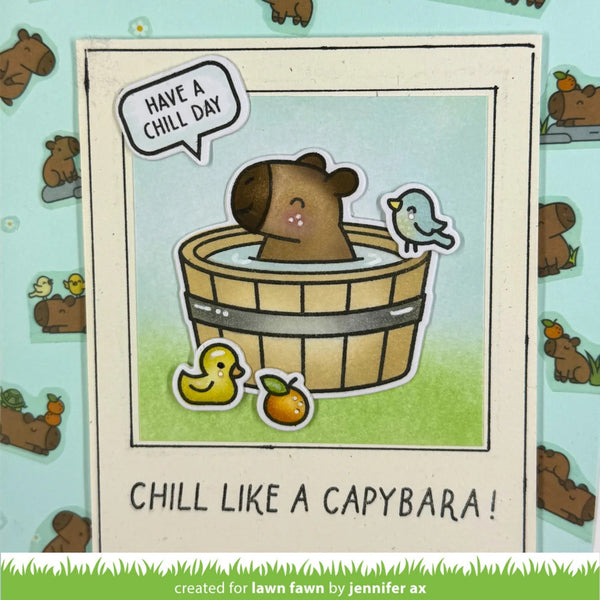 A handmade greeting card featuring a cute cartoon capybara in a wooden bath tub with a small bird and duck, with the text 'HAVE A CHILL DAY!' and 'CHILL LIKE A CAPYBARA!'. The card is displayed on a light blue patterned background.