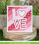 Card with 'LOVE' and heart design on a pink and white patterned background, placed on a wooden block with a natural setting.