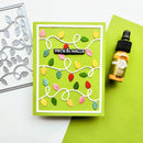 A handmade greeting card with a green background, white border, and colorful paper Christmas light garland, styled on a white surface with a green paper sheet and a small amber dropper bottle. A metal die-cut template is partially visible on the left.