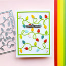 A handmade greeting card featuring colorful Christmas string lights on a white background with a green border, displayed next to a metal die-cutting template on a pink background.