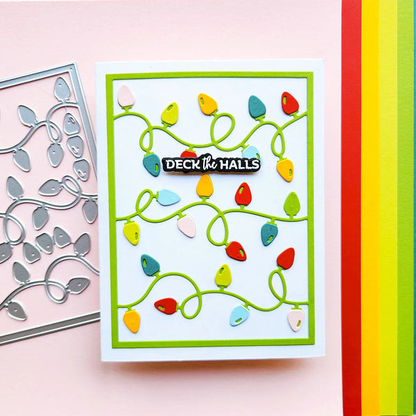 A handmade greeting card featuring colorful Christmas string lights on a white background with a green border, displayed next to a metal die-cutting template on a pink background.