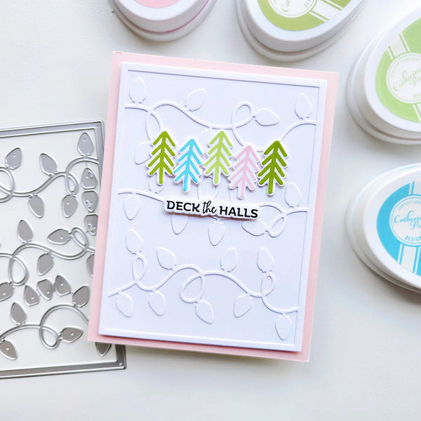 A handmade greeting card with a white embossed background, a pink border, and four colorful pine trees above the words 'DECK THE HALLS'. The card is styled on a white surface with crafting tools, including a metal die and small ink pads.