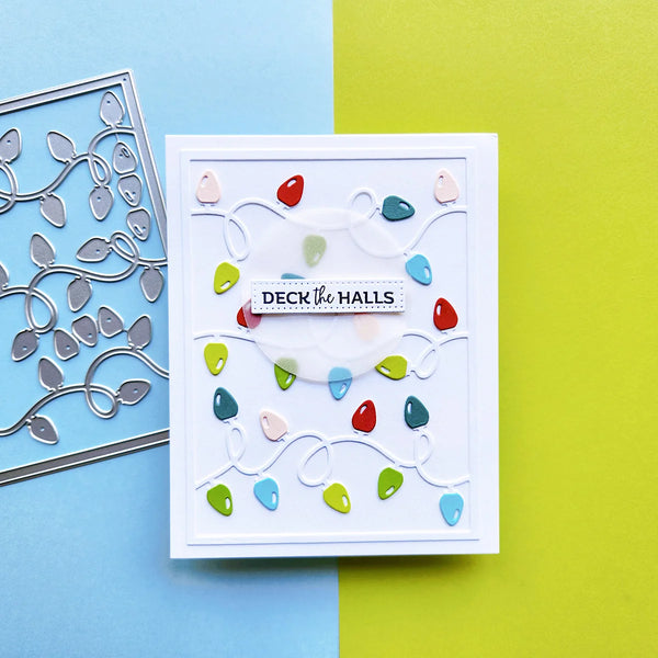 A handmade layered card featuring colorful Christmas string lights and a central banner reading 'DECK THE HALLS', styled on a split blue and yellow background with a metal die cut sheet partially visible on the left.