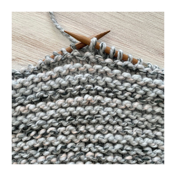 Close-up of a knitted fabric swatch in a variegated gray, white, and soft pink yarn, held on wooden knitting needles over a light wood background.