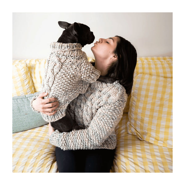 A woman in a chunky knit beige sweater holds a small black dog wearing a matching cable-knit sweater while sitting on a yellow-and-white checkered couch.