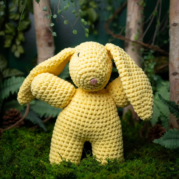 Yellow crochet bunny toy in a natural setting with plants and grass.